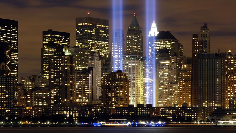Kuvassa on öinen Manhattanin siluetti, jossa on kerrostaloja ja niiden ikkunoista paistaa valoja. Talojen keskeltä nousee kaksi suurta ylöspäin kohdistettua valopylvästä.