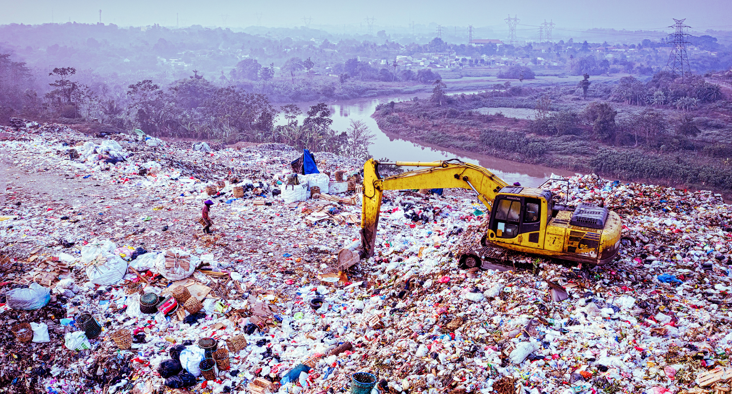 Laaja maisema, jonka etualalla on suuri kaatopaikka-alue. Roskaläjien päällä on keltainen kaivinkone. Taustalla näkyy metsäisen vyöhykkeen läpi kiemurteleva joki ja kaupunkimaista aluetta.