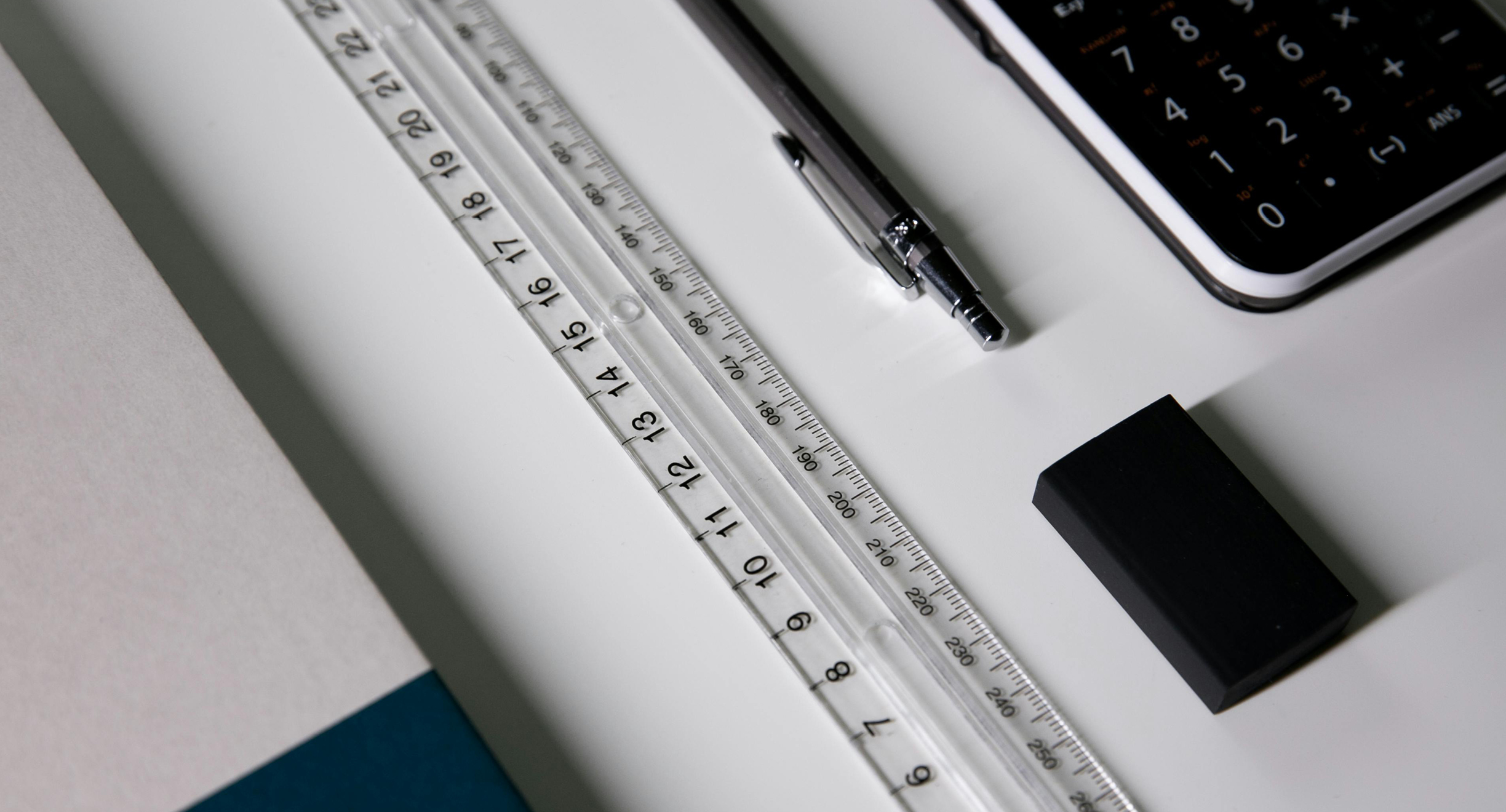 Valkoisella pöydällä on muutamia toimistotarvikkeita: muistikirja, viivotin, kynä ja laskin.