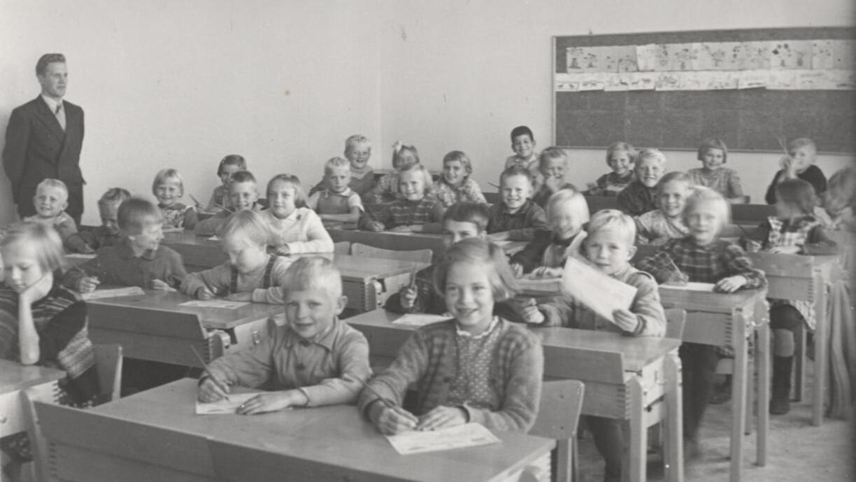 Mustavalkoinen kuva, jossa alakoululaisia istuu pulpeteissa koululuokassa.