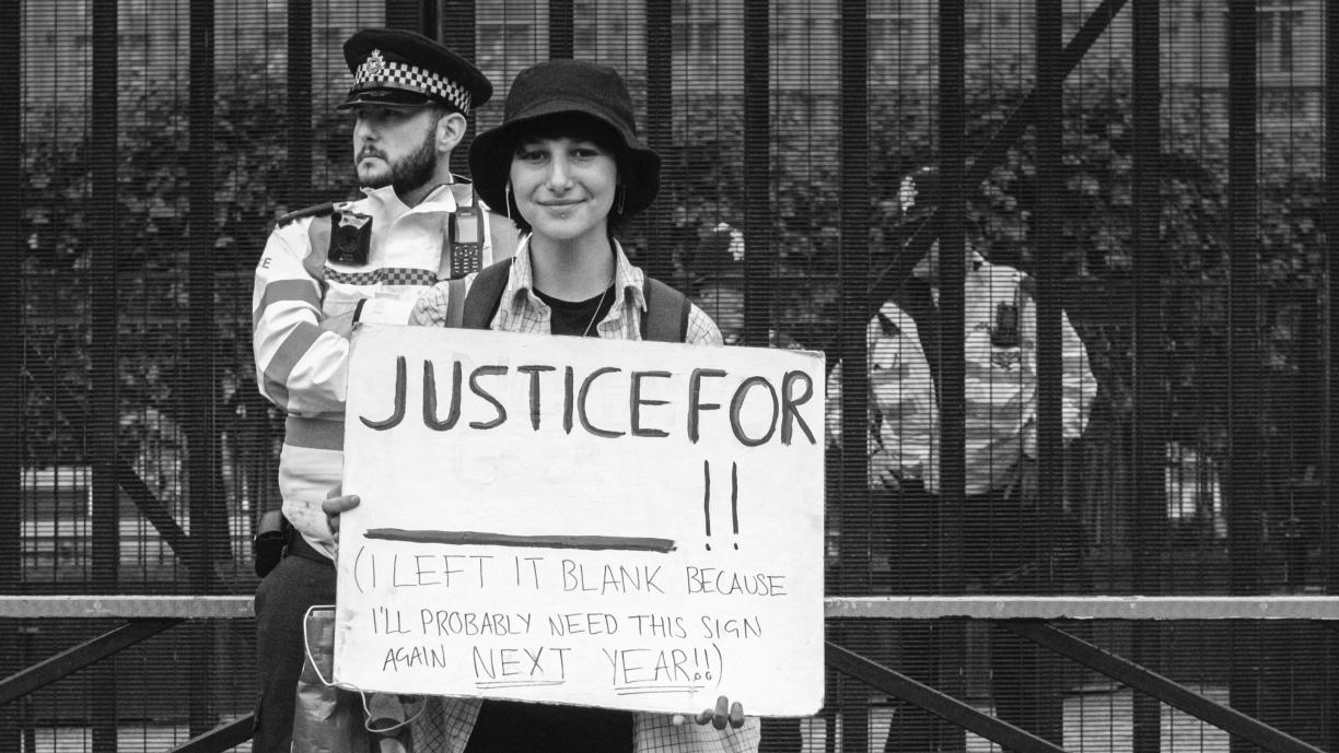 Henkilö seisoo kadulla korkean aidan edessä ja pitää käsissään kylttiä, jossa lukee "Justice for - ! (I left blank because I'll probably need this sign again next year!)". Henkilön takana seisoo poliisi ympäristöä tarkkailemassa.