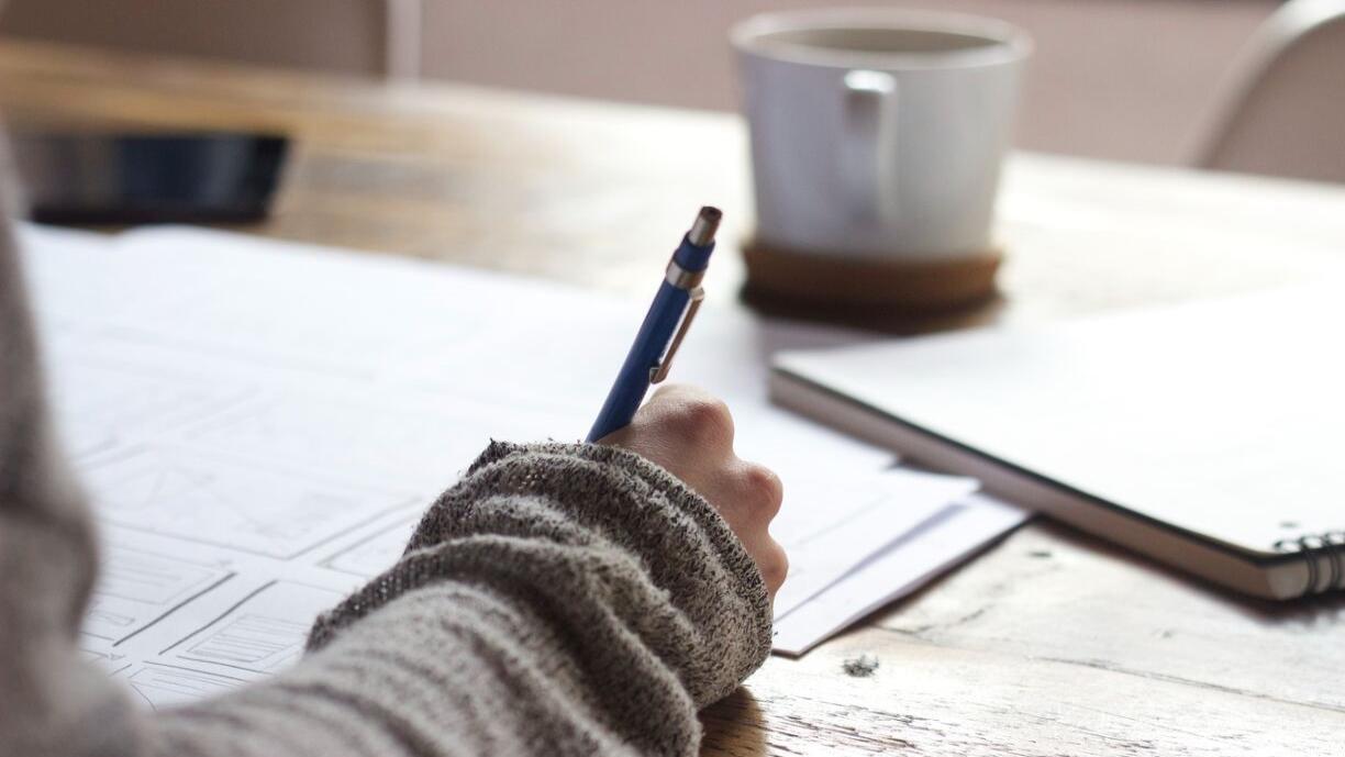 Valokuva, jossa näkyy papereihin kynällä kirjoittava käsi ja kahvikuppi pöydällä.