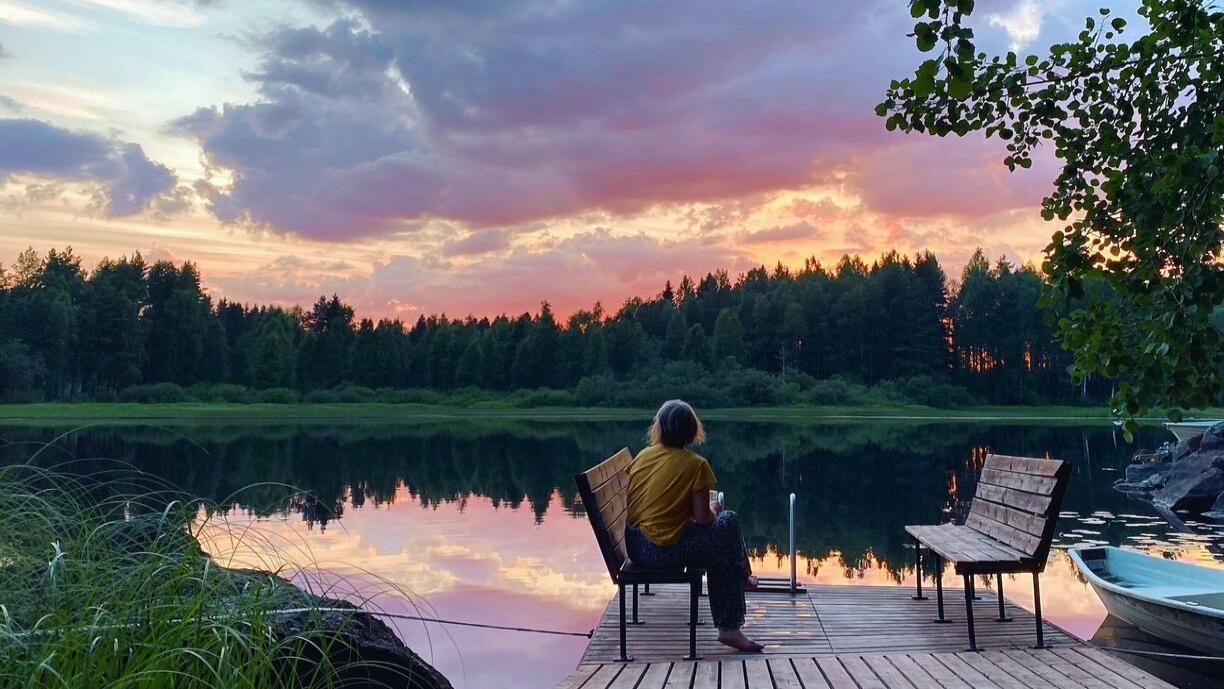 Nainen istuu laiturilla ja katsoo järvelle, jonka toisella rannalla näkyy havumetsää.