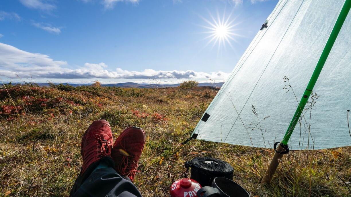 Valokuva, jossa nkyy maastossa istuvan retkeilijän jalat Kilpisjärvellä. Aurinko paistaa taivaalla.