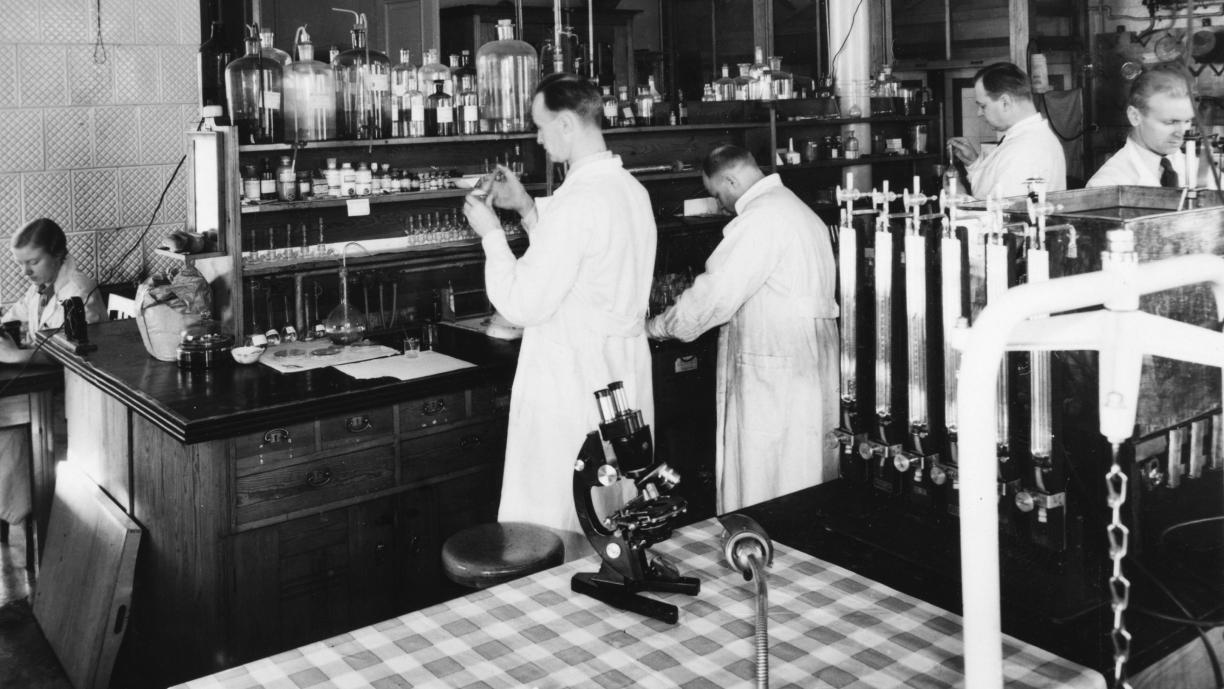 Mustavalkoinen valokuva, jossa viisi henkilöä työskentelee valkoiset takit päällä erilaisten pullojen, laitteiden ja putkistojen ääressä.