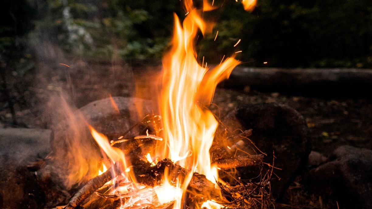 Oranssina roihuava nuotio kivien keskellä tummassa metsämaisemassa.