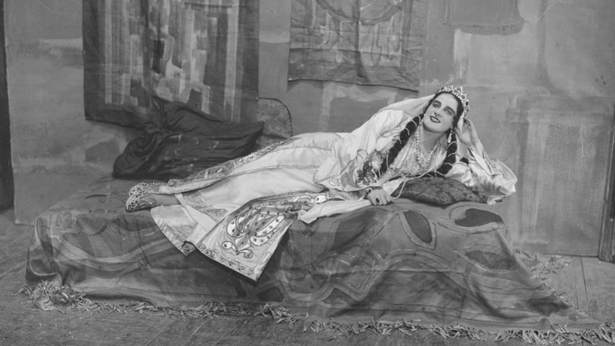 Mustavalkoinen valokuva, jossa valkoiseen esiintymisasuun pukeutunut nainen makaa vuoteella.