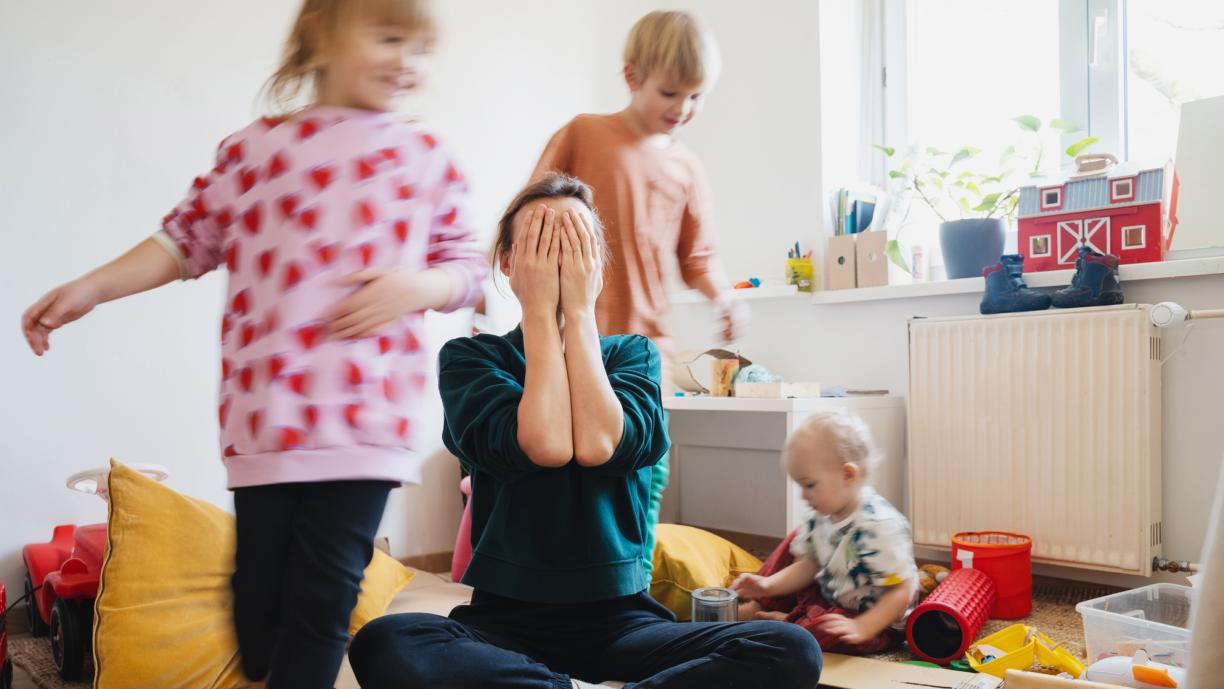Sisäkuva huoneesta. Naisoletettu istuu lattialla ja peittää kasvonsa käsillään. Hänen ympärillään kaksi lasta juoksee ja yksi istuu lattialla. Lattialla ja tasoilla on erilaisia leluja ja muita esineitä.