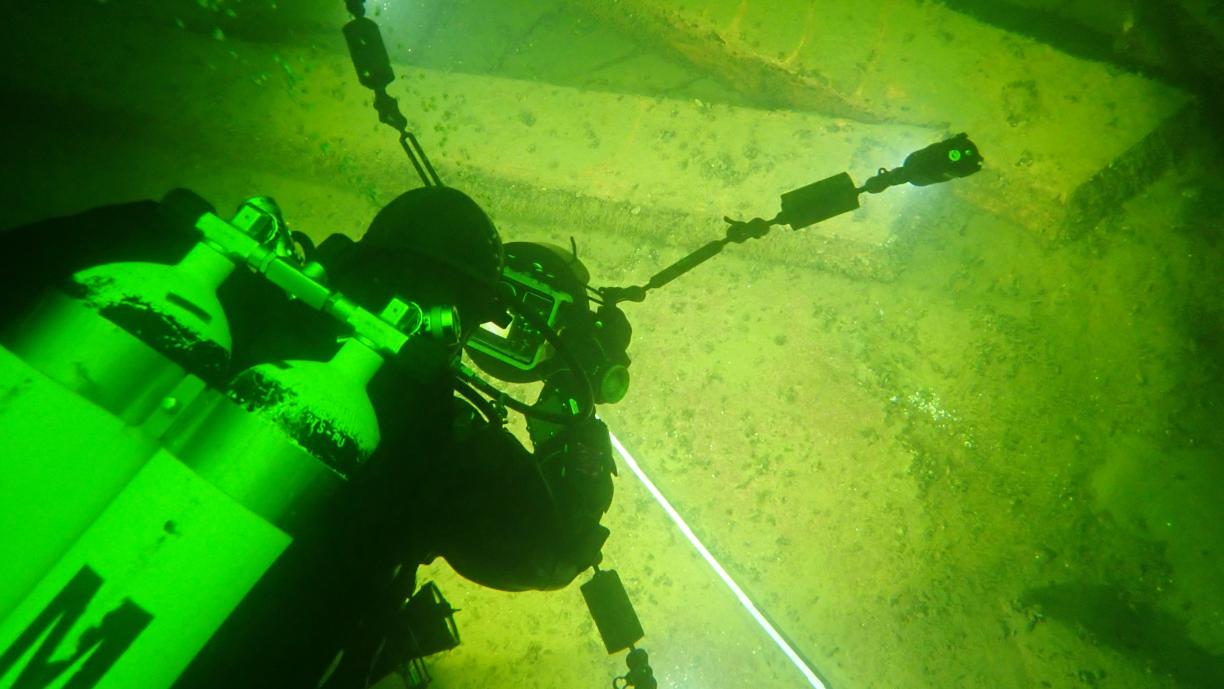 Sukeltaja happipulloineen kuvaa veden alla näkyviä suuria palkin näköisiä elementtejä. Maisema on vihertävä ja kamera valaisee kuvattavaa kohdetta.