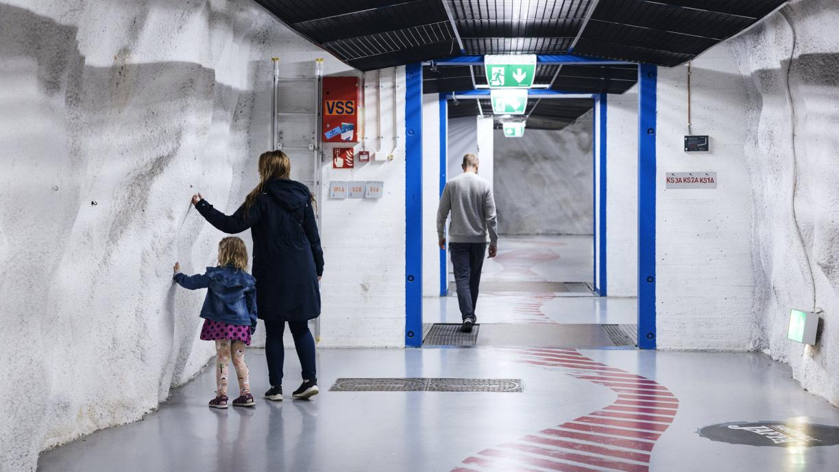 Ikkunaton vaaleaseinäinen leveä käytävä, jossa kävelee muutamia ihmisiä. Seinillä on joitakin laitteita johtoineen.