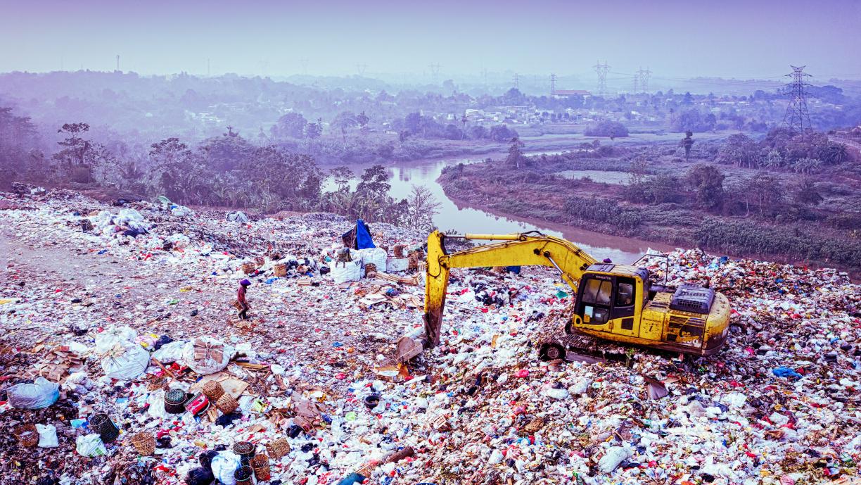 Laaja maisema, jonka etualalla on suuri kaatopaikka-alue. Roskaläjien päällä on keltainen kaivinkone. Taustalla näkyy metsäisen vyöhykkeen läpi kiemurteleva joki ja kaupunkimaista aluetta.
