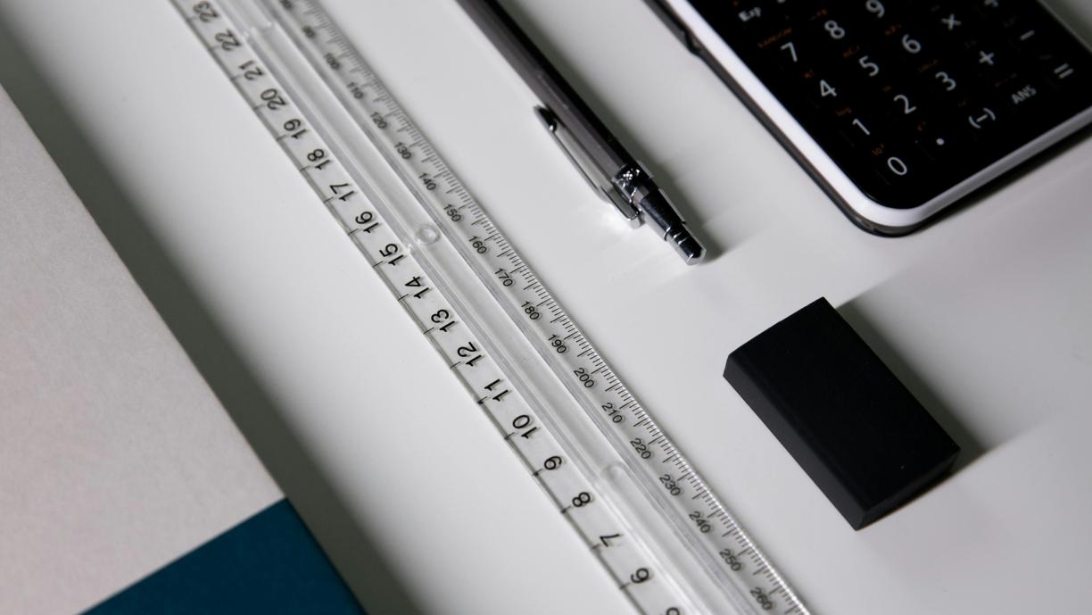 Valkoisella pöydällä on muutamia toimistotarvikkeita: muistikirja, viivotin, kynä ja laskin.