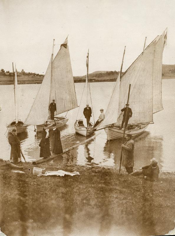 Mustavalkoisessa kuvassa on neljä pientä venettä rannan tuntumassa. Veneiden kansilla ja rannassa seisoo siististi pukeutuneita miehiä ja naisia.