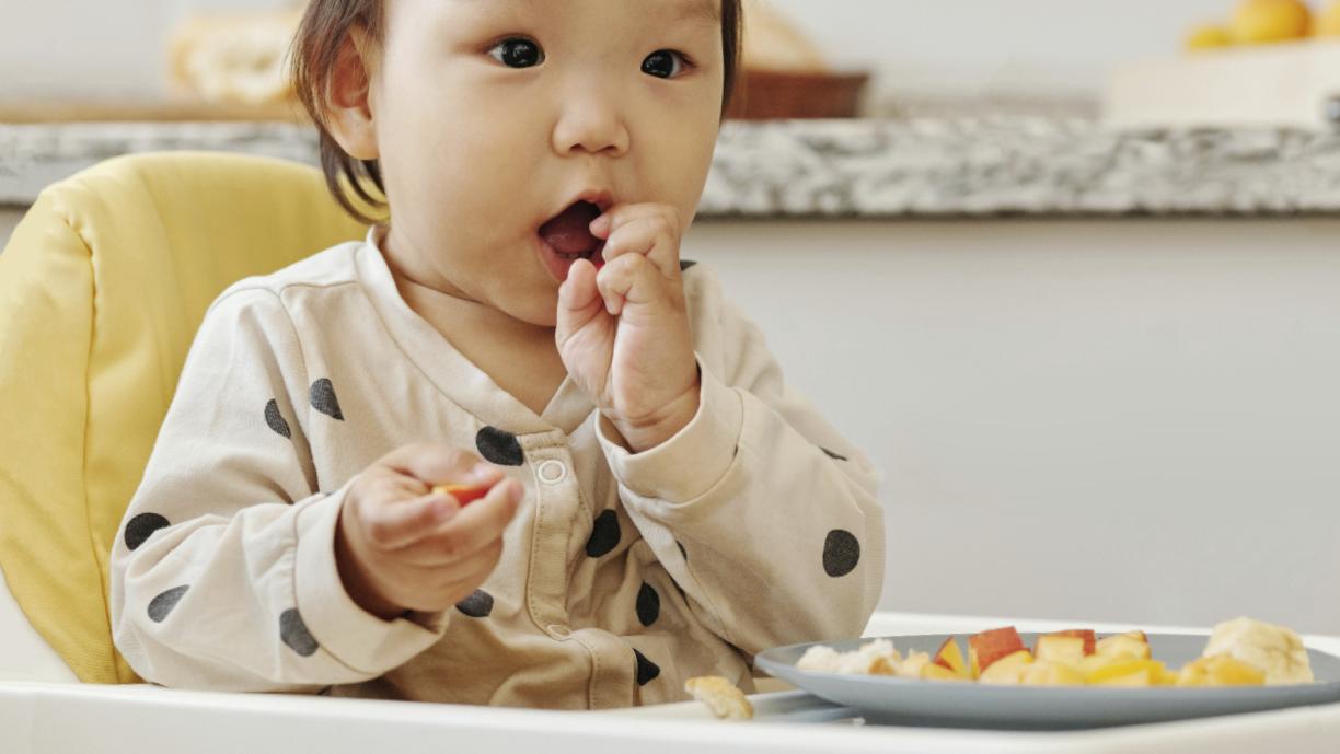 Pieni lapsi istuu syöttötuolissa. Hän on syömässä pilkottuja hedelmiä, joita on hänen edessään lautasella.