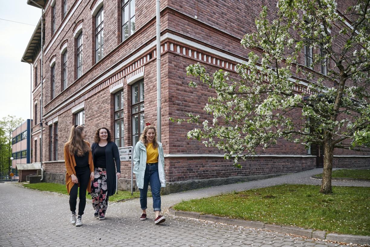 Kolme nuorta naista kävelee punatiilisen rakennuksen edessä.