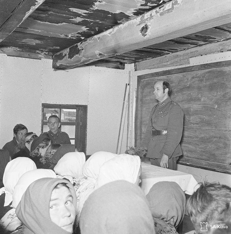 Mustavalkoinen valokuva huoneesta, jossa joukko ihmisiä katsoo liitutaulun edessä seisovaa asepukuista miestä.