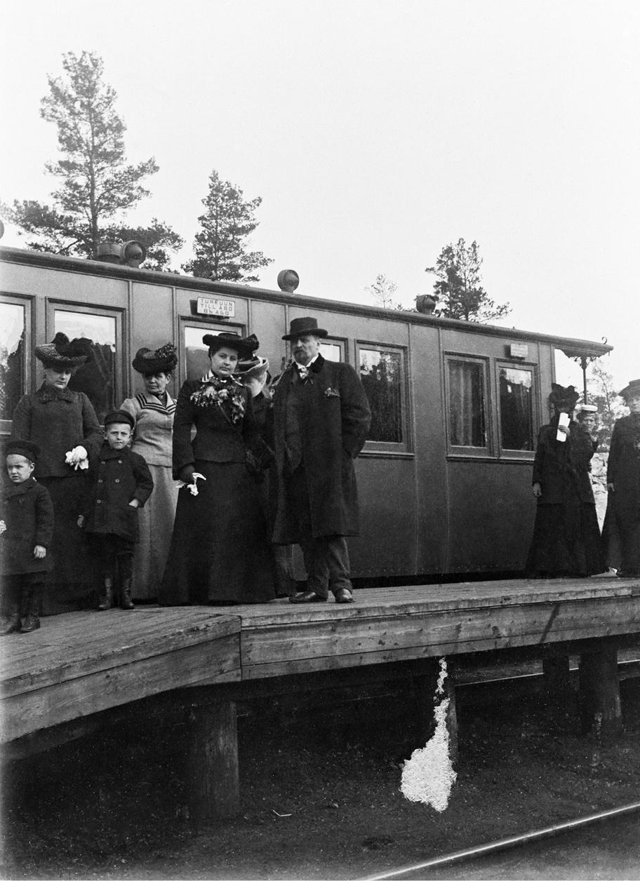Mustavalkoinen valokuva, jossa seisoo ihmisiä puisella junalaiturilla. Taustalla näkyy junan vaunu, jonka oven päällä on kyltti "Turkuun, Till Åbo".