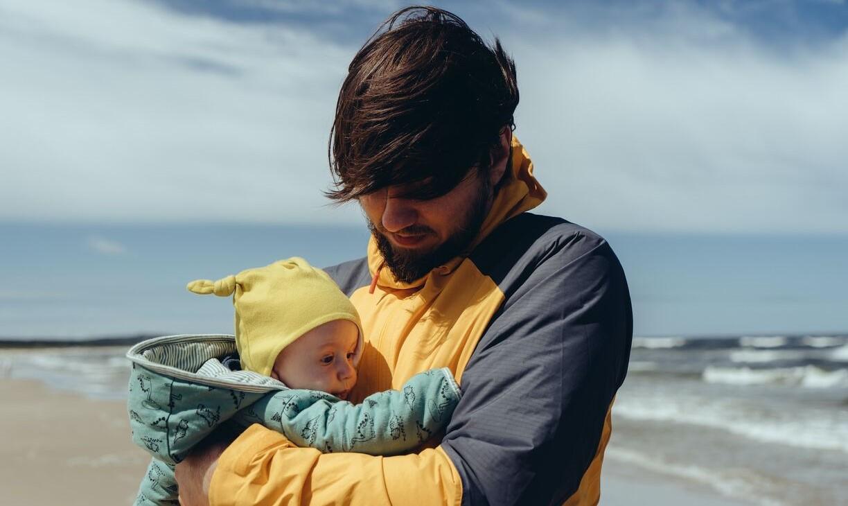 Mies pitelee lasta sylissään tuulisella rannalla.