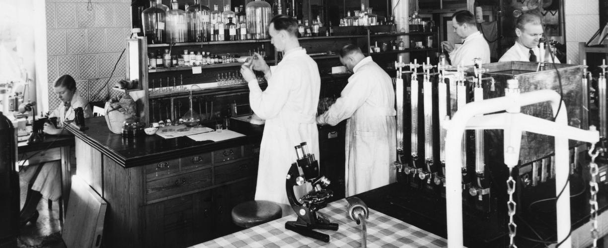 Mustavalkoinen valokuva, jossa viisi henkilöä työskentelee valkoiset takit päällä erilaisten pullojen, laitteiden ja putkistojen ääressä.