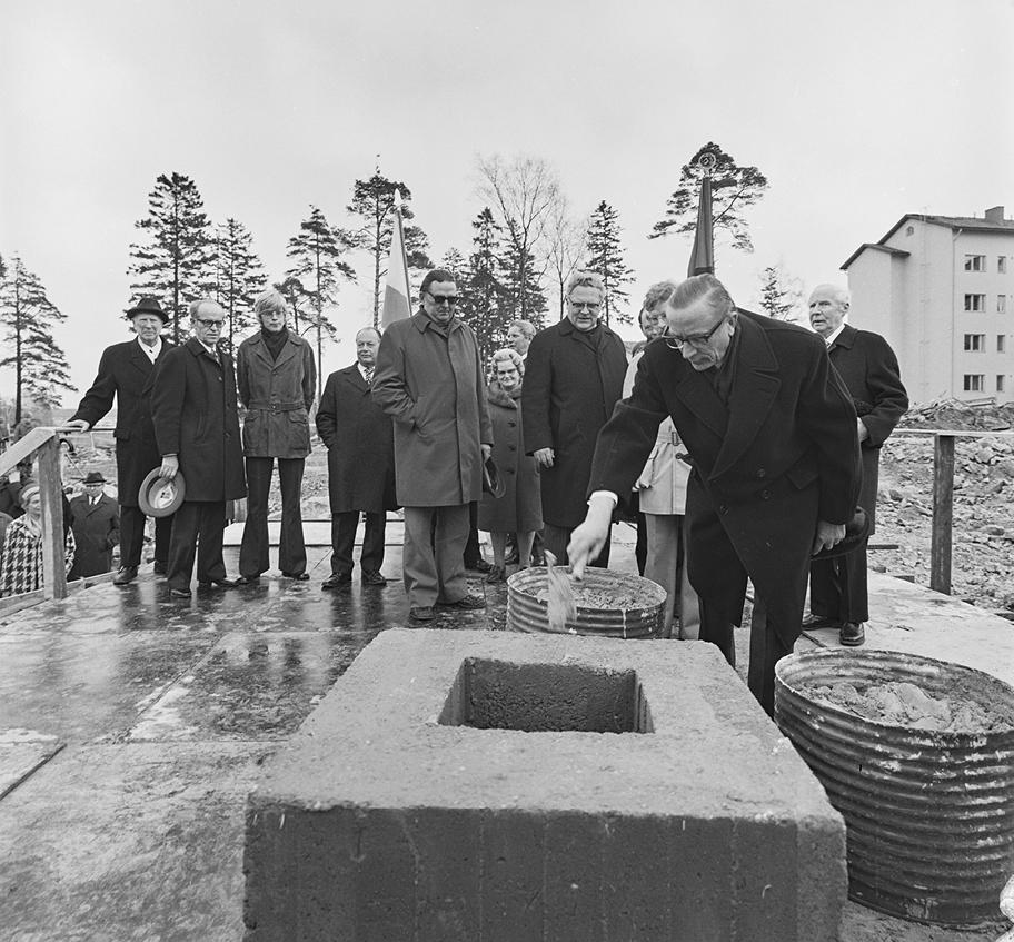 Mustavalkoinen valokuva, jossa viitisentoista ihmistä seisoo ulkona rakennustyömaan keskellä olevalla korokkeella. Etualalla oleva mies kauhoo ämpäristä sementtiä suureen kehikoon.