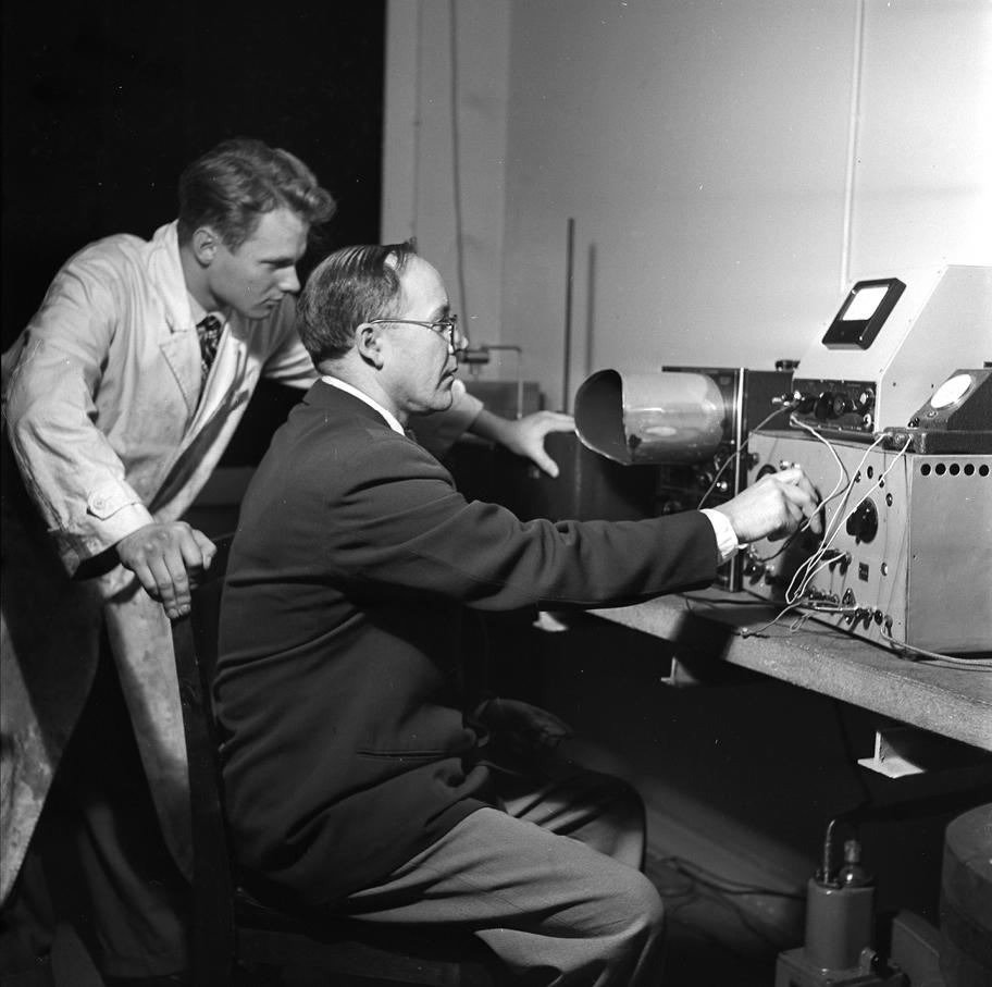Mustavalkoinen valokuva, jossa kaksi miestä työskentelee pöydällä olevan kookkaan laitteen ääressä.
