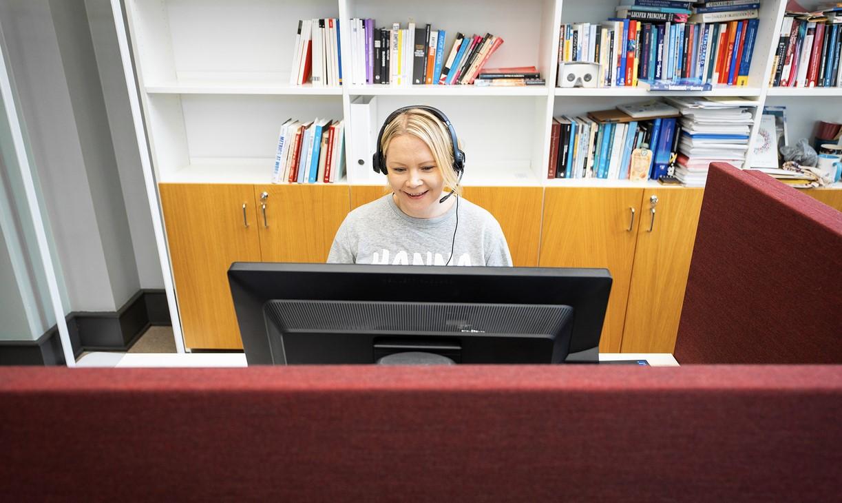 Nainen istuu tietokoneen ääressä kuulokkeet päässään ja näyttää puhuvan mikrofoniin.