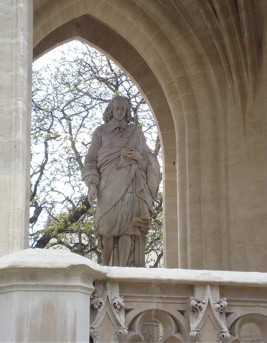 Vaaleasta kivestä veistetty patsas miehestä, jolla on pitkät kiharat hiukset ja koristeellinen asu. Patsas sijaitsee korkean holvikaaren alla, etualalla on koristeellinen aita.