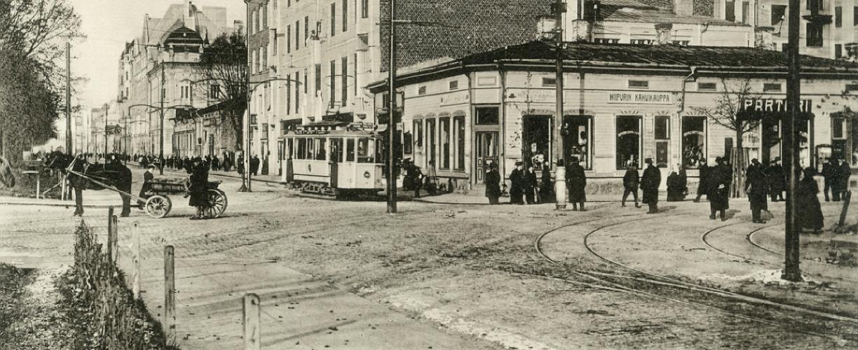 Mustavalkoinen valokuva kadunkulmasta. Katu on melko avara, ja sillä kävelee ihmisiä, ajaa raitiovaunu ja kulkee hevoskärryt. Yksi rakennuksista on yksikerroksinen liikerakennus, muut kolmi- tai nelikerroksisia kivirakennuksia. Kadun reunassa kulkee sähkölinjoja ja sen toisella puolella näkyy puita.