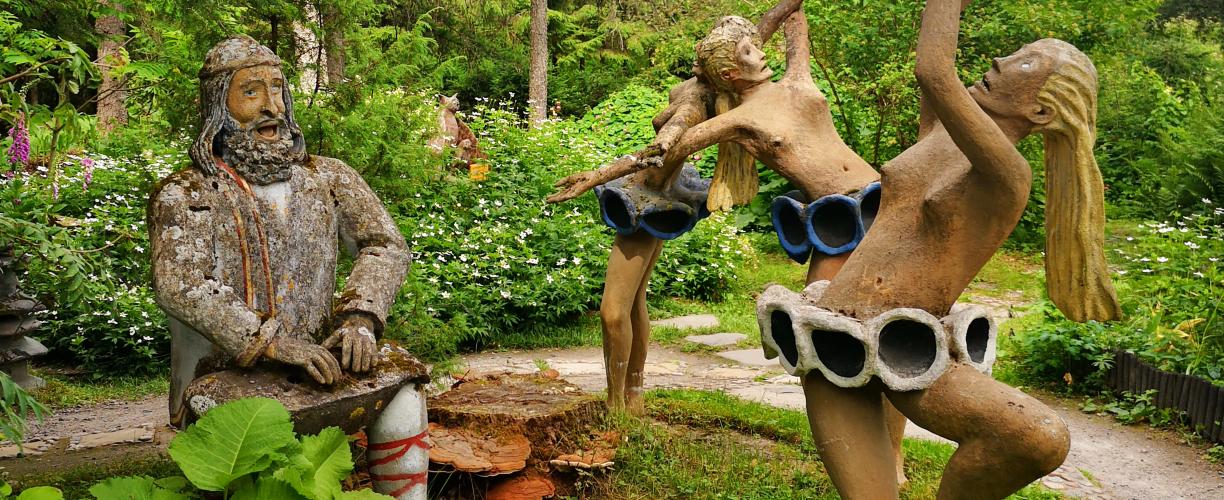 Neljä patsasta vihreässä metsä- ja puutarhamaisemassa. Yksi patsaista näyttää Väinämöiseltä kanteleineen ja perinneasuineen, kolme patsasta tanssii. Tanssijoilla on päällään vain erikoisen näköiset lyhyet hameet.