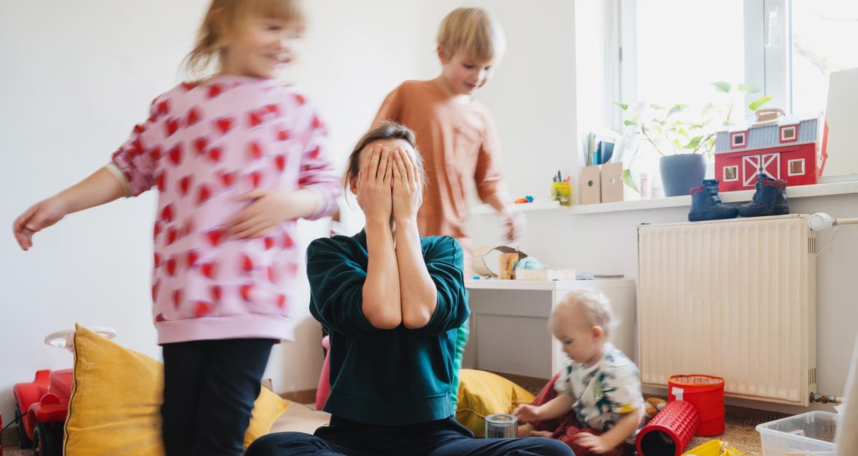 Sisäkuva huoneesta. Naisoletettu istuu lattialla ja peittää kasvonsa käsillään. Hänen ympärillään kaksi lasta juoksee ja yksi istuu lattialla. Lattialla ja tasoilla on erilaisia leluja ja muita esineitä.