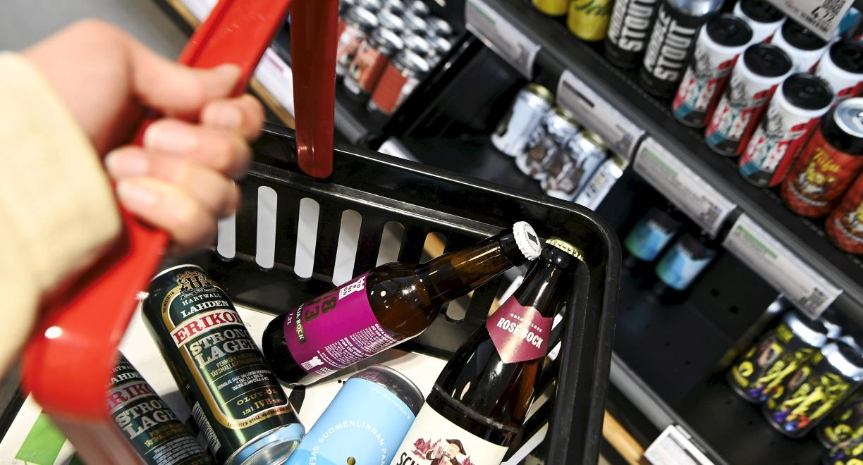 Ostoskori, jossa on kolme tölkkiä ja kaksi pulloa erimerkkisiä olluita. Taustalla näkyy hylly, jossa on runsaasti lisää alkoholijuomia.