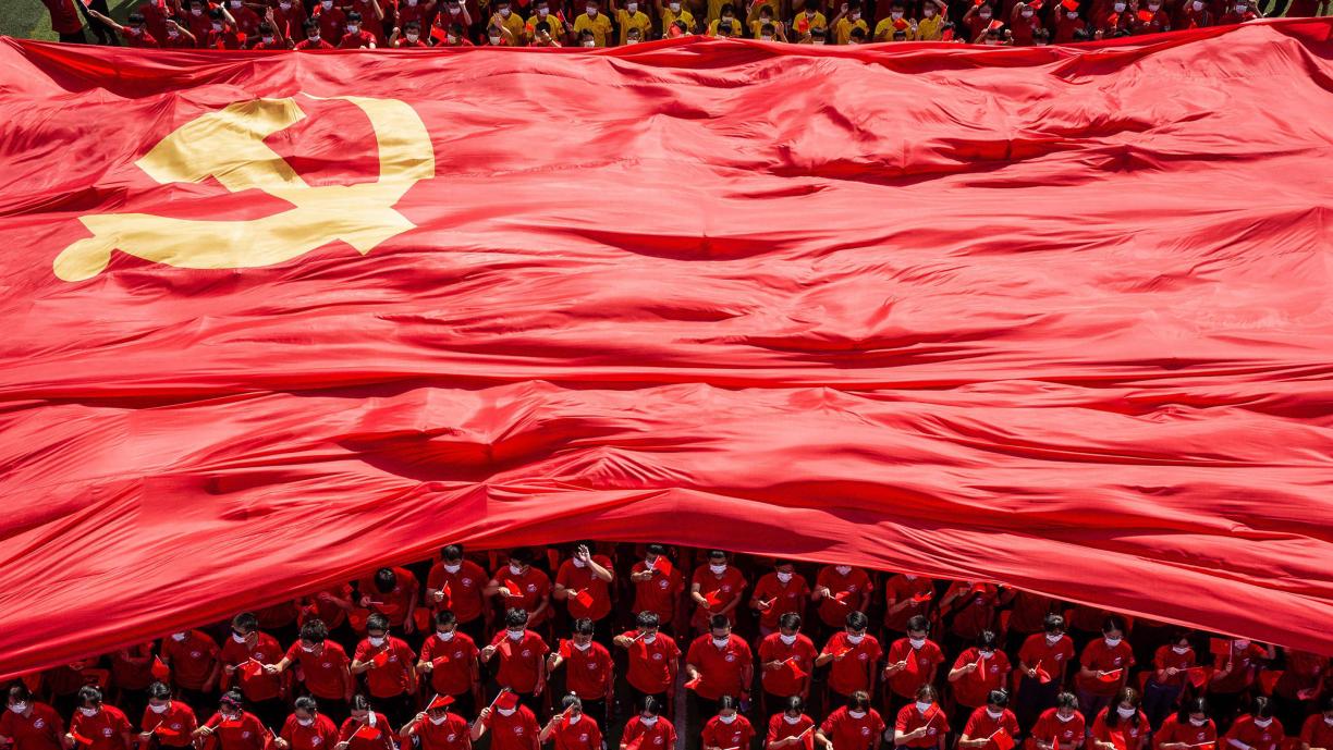 Punaisiin pukeutuneita nuoria seisoo katsomossa. Heidän päälleen on avautumassa suuri kankainen Kiinan lippu.