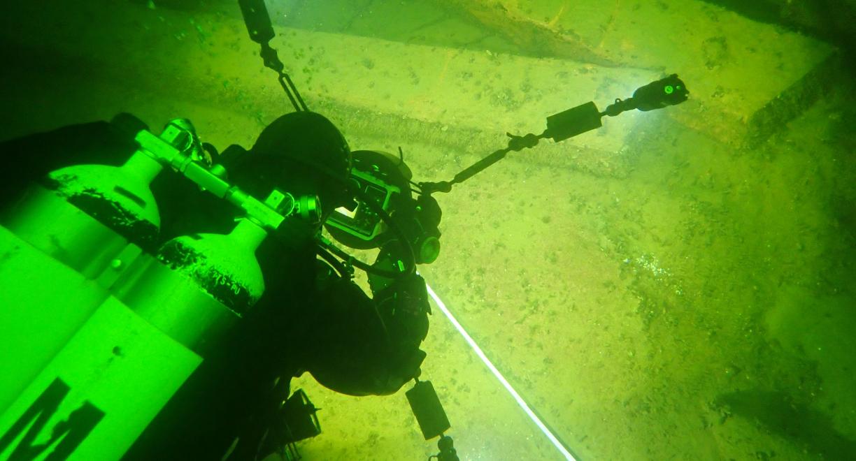 Sukeltaja happipulloineen kuvaa veden alla näkyviä suuria palkin näköisiä elementtejä. Maisema on vihertävä ja kamera valaisee kuvattavaa kohdetta.