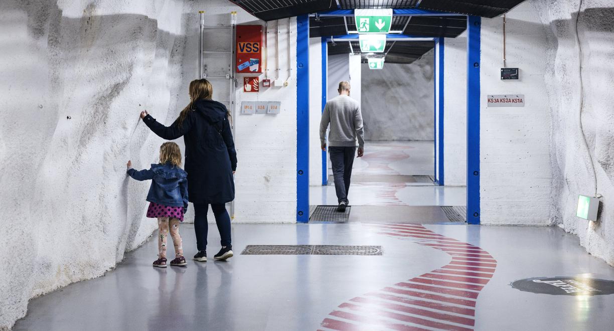 Ikkunaton vaaleaseinäinen leveä käytävä, jossa kävelee muutamia ihmisiä. Seinillä on joitakin laitteita johtoineen.