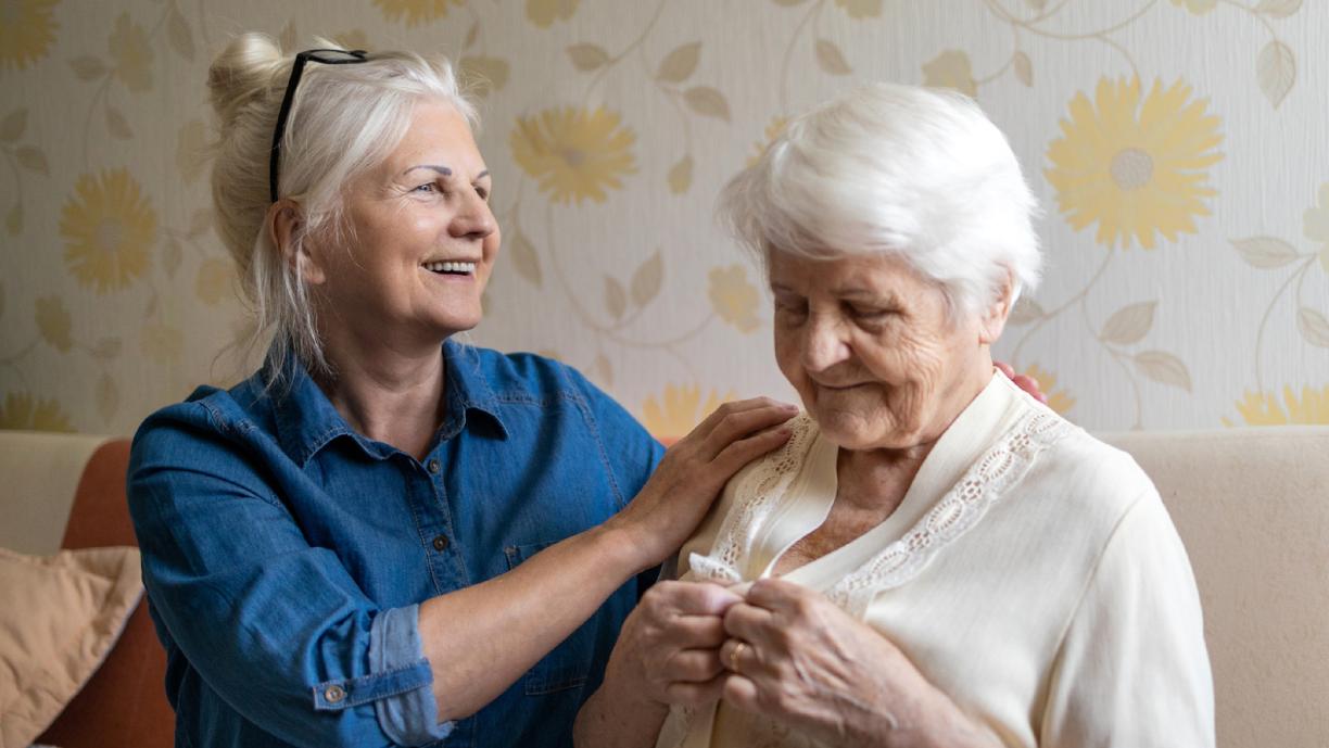 Kaksi naisoletettua istuu sohvalla. Molemmilla on harmaat hiukset. Toinen, hieman nuoremman näköinen, pitää toista hartioista ja toinen napittaa puseroaan.