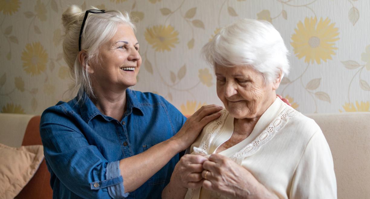 Kaksi naisoletettua istuu sohvalla. Molemmilla on harmaat hiukset. Toinen, hieman nuoremman näköinen, pitää toista hartioista ja toinen napittaa puseroaan.