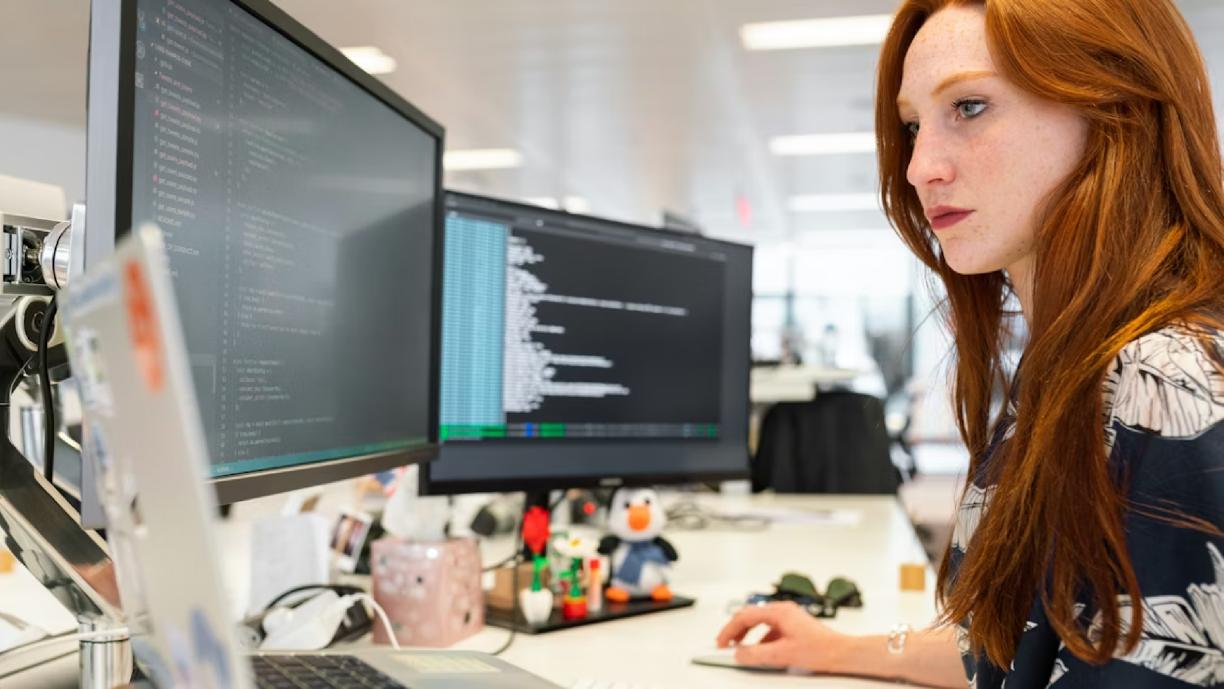 Naisoletettu istuu työpöydän ääressä ja katsoo tietokoneen näyttöä. Pöydällä on kaksi näyttöä ja yksi kannettava tietokone sekä muuta esineistöä.