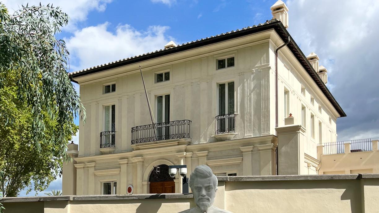 Vaalea kivitalo. Talon edessä on aita ja aidan edessä vaaleasta kivestä tehty miehen rintakuva. Talon vieressä on tuuhea lehtipuu.