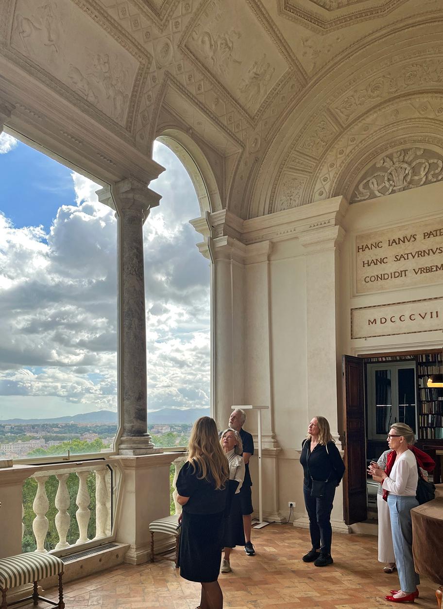 Kuusi ihmistä seisoo vaaleasta kivestä tehdyn rakennuksen avaralla, korkealla parvekkeella. Katossa on kaaria ja reliefejä. Parvekkeelta avautuu avara näkymä maisemaan, jossa on rakennuksia, metsää ja kaukana matalia vuoria.