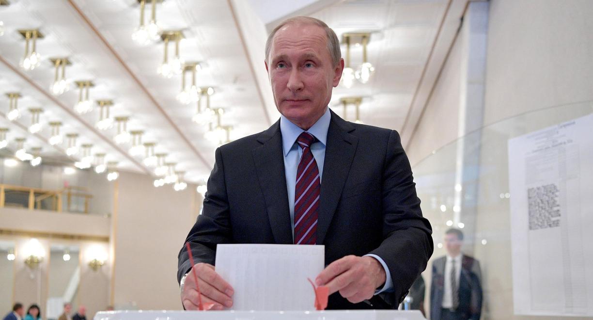 Pukuun ja solmioon pukeutunut mies asettamassa valkoista paperia suureen laatikkoon.