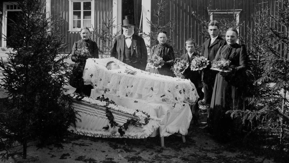 Mustavalkoinen valokuva, jossa kuusi ihmistä seisoo mustissa vaatteissa talon pihamaalla avoimen arkun edessä. Monilla heistä on kukkakimput käsissään. Arkku on vuorattu ja päällystetty valkoisella kankaalla ja arkussa makaa nuori ihminen. 