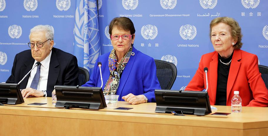 Valokuva kolmesta hahmosta, jotka istuvat vierekkäin kokouspöydän ja mikrofonien äärellä. Keskimmäisenä on tummahiuksinen, silmälasipäinen nainen, joka katsoo kameraan. Taustalla on sininen seinä, jolla on YK:n tunnus ja nimi monella eri kielellä.