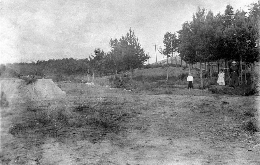 Mustavalkoinen hieman epätarkka valokuva, jossa nainen ja kaksi lasta seisoo hiekkaisen aukion laidalla. Tautalla on kumpuilevaa metsää ja puuaita. Aukiolla näkyy suuri vaalea koroke, jonka päällä on seppele nauhoineen.