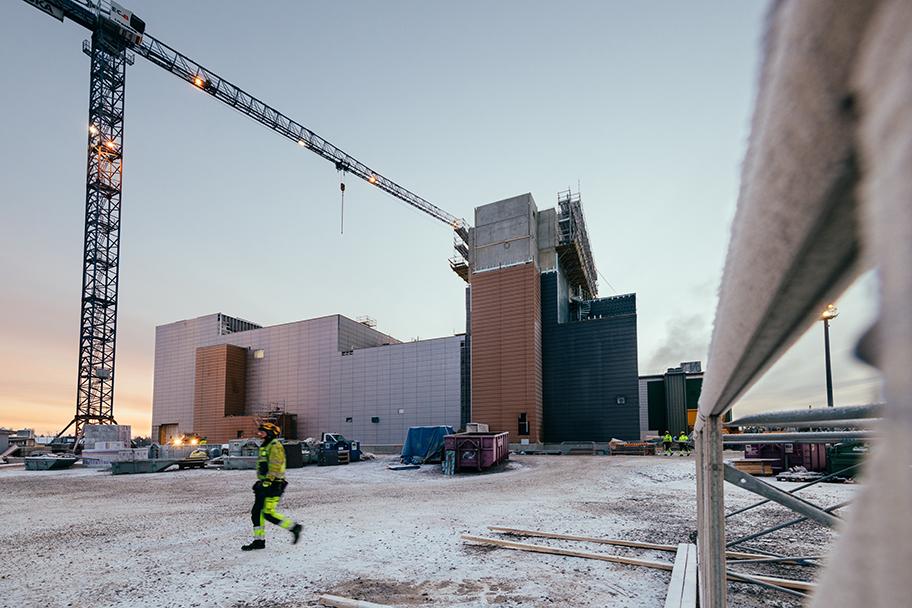 Laaja näkymä teollisuuslaitoksesta ja sen pihasta kevyessä hämärässä. Pihalla näkyy kolme työmiestä kirkkaissa työasuissa ja kypärissä.