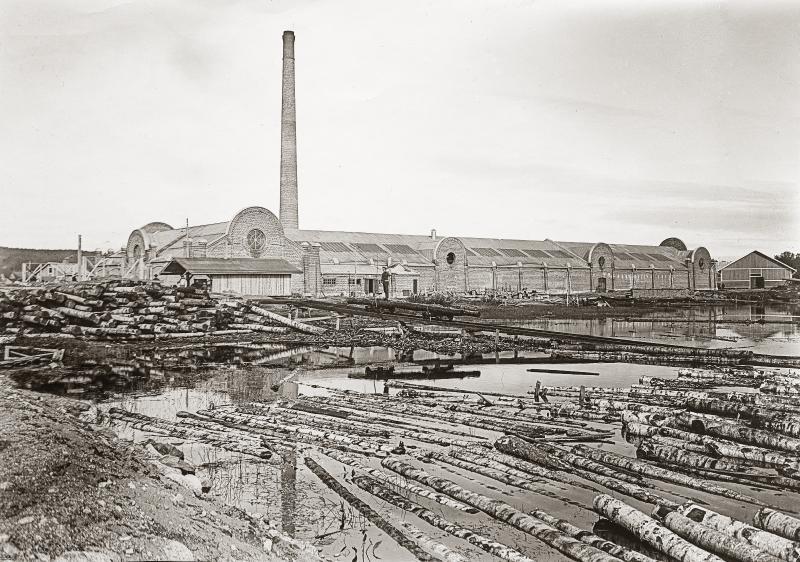 Mustavalkoisessa kuvassa on järven rannalla sijaitseva tiilestä rakennettu tehdasrakennus, jolla on korkea piippu. Kuvan etualalla on järvessä koivutukkeja.