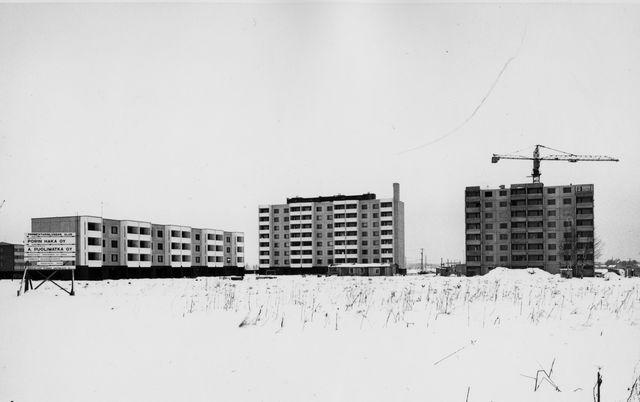 Mustavalkoisessa kuvassa on vierekkäin kolme betonielementeistä rakenteilla olevaa taloa talvisessa maisemassa. Oikeanpuoleisen talon päällä on nostokurki.