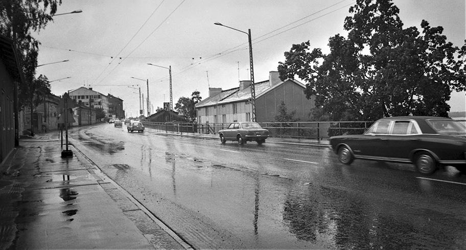 Mustavalkoinen valokuva sateisesta kaupunkimaisemasta, jossa useat rakennukset ja muutamat puut reunustavat leveää tietä. Tiellä ajaa vajaa kymmenen autoa.