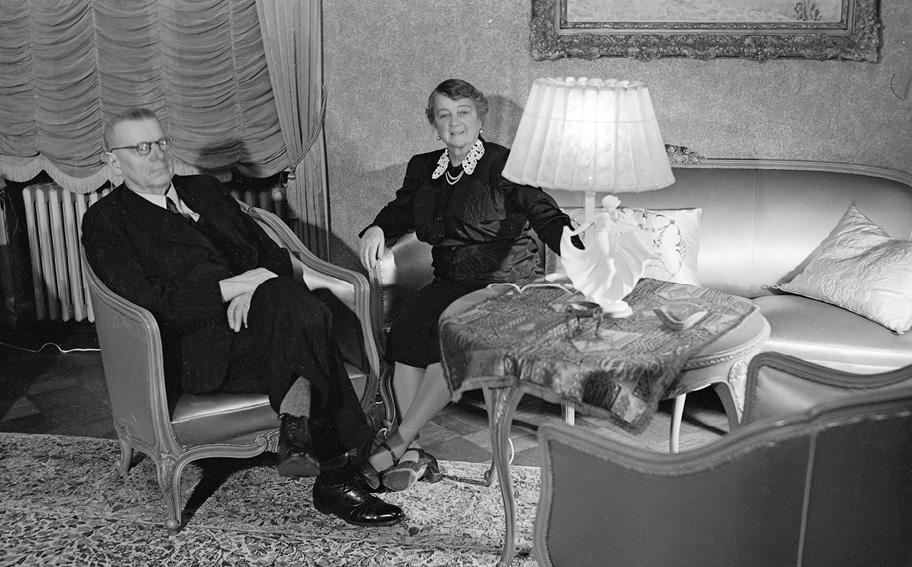 Mustavalkoinen valokuva tyylikkäästi sisustetusta huoneesta. Mies tummassa puvussa istuu nojatuolilla, nainen tummassa asussa istuu vieressä sohvalla.