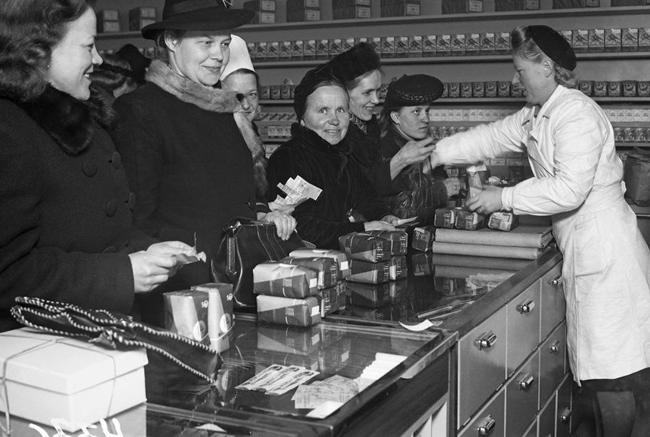 Mustavalkoinen valokuva kaupasta. Tiskillä on kuusi rouvaa odottamassa vuoroaan, yksi myyjätäs palvelee heitä. Kaikilla asiakkailla on tiskillä odottamassa samannäköisiä paperipäällysteisiä pakkauksia.