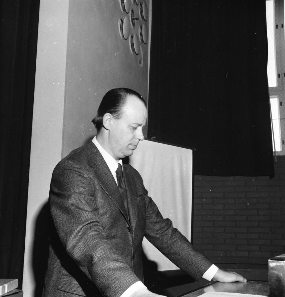 Mustavalkoinen kuva, jossa pukuun pukeutunut mies nojaa puhujan korokkeeseen.